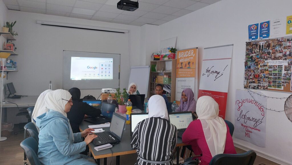 In einem großen Raum sitzen acht Frauen an einem großen Tisch und jede Frau hat einen Laptop vor sich geöffnet. An einem Whiteboard im Hintergrund ist aktuell die Startseite der Google Suchmaschine sichtbar.