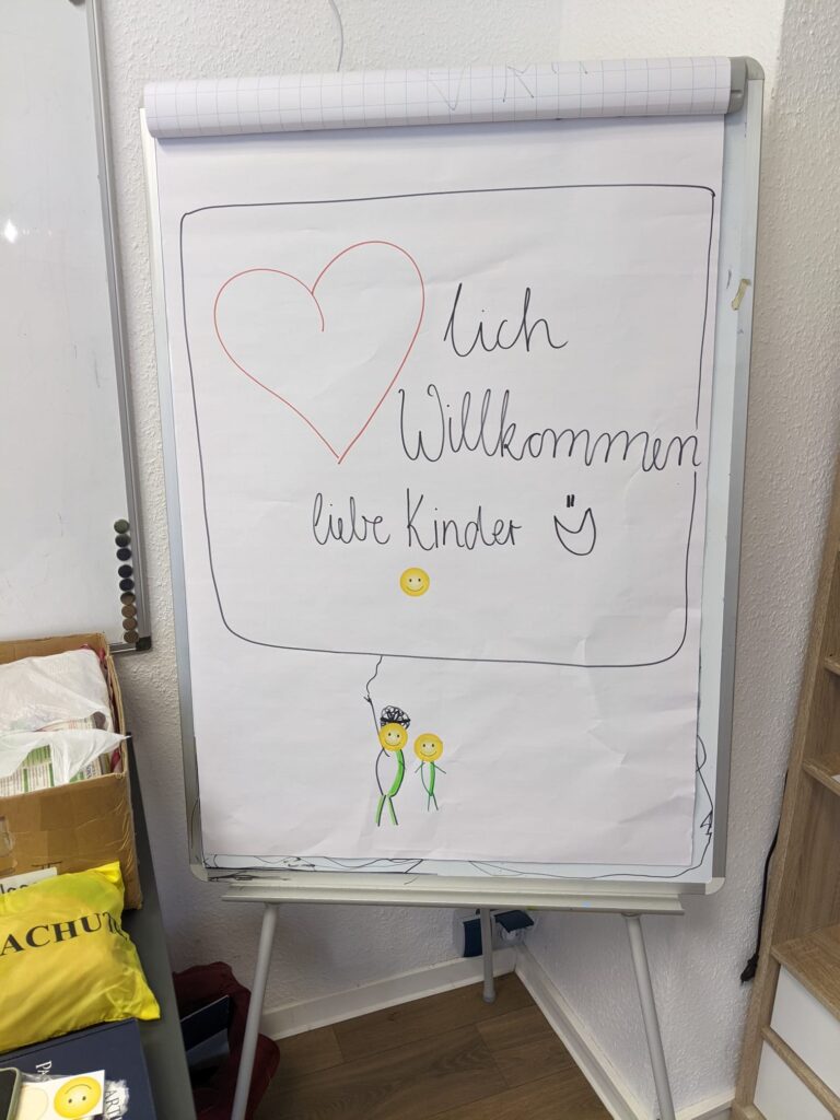 Auf einem großen weißen Flipchart-Papier wurde liebevoll "Herzlich Willkommen liebe Kinder" geschrieben, wobei das Herz im Wort Herzlich ein gezeichnetes Herz ist. Hinzukommend sind noch kleine Zeichnungen drauf, wie Smilies und zwei Strichzeichnungen von Menschen.