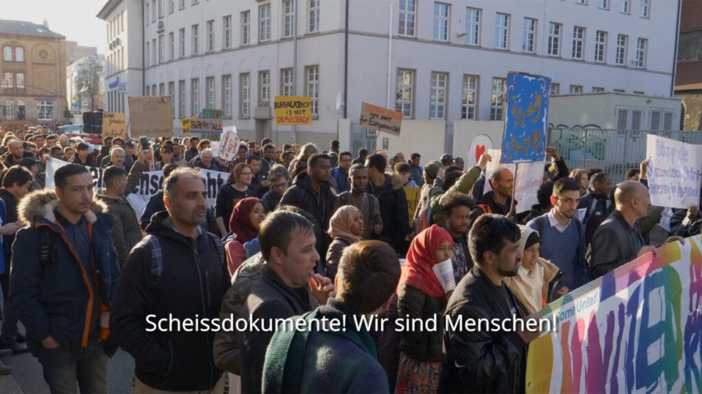 Standbild von einer Demonstrations-Szene: Viele Menschen tragen kleine Schilder und großformatige Transparente gegen Bürokratie im Asylverfahren. Im Video-Untertitel steht: Scheissdokumente! Wir sind Menschen!
