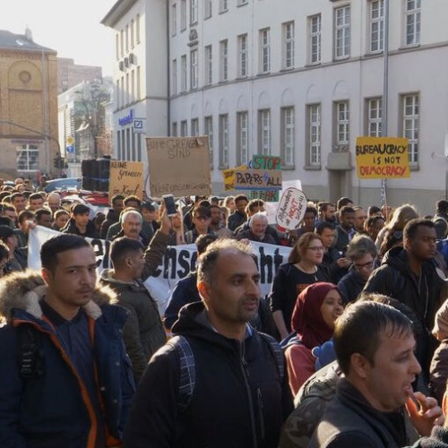 Standbild von einer Demonstrations-Szene: Viele Menschen tragen kleine Schilder und großformatige Transparente gegen Bürokratie im Asylverfahren.