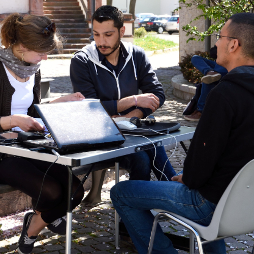Draußen im Schatten sitzen drei Personen mit einem Laptop an einem Tisch.