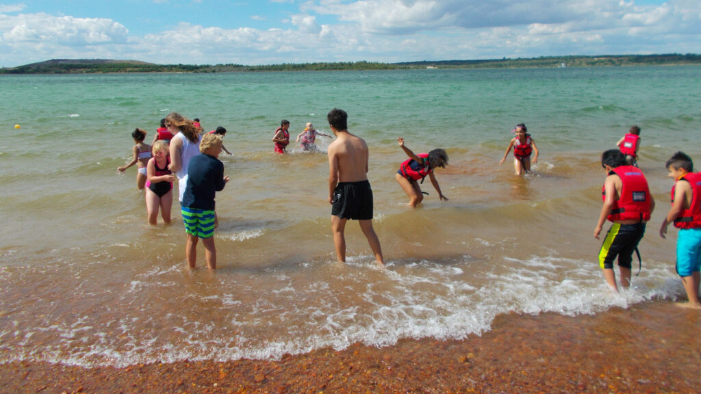 Circa 15 Kinder, davon die Hälfte in roten Schwimmwesten spielen im Wasser am Ufer eines Sees, der grün-blau leuchtet und kupferfarbene Kieselsteine hat. Es wirkt sehr windig und kalt. Im fernen HIntergrund sind Bäume und ein kleiner Hügel.