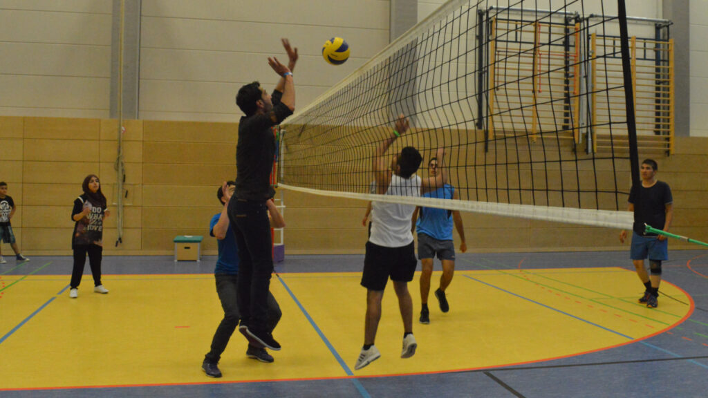 Zwei Teams stehen sich direkt an einem Volleyballnetz gegenüber. Ein Spieler auf der linken Seite versucht gerade am Netz im Sprung den kommenden gelb-blauen Volleyball zu blocken. Sein Gegenspieler auf der anderen Seite ist kurz vor der Landung seines Sprungs. Auf jeder Seite sind zwei weitere Spieler zu sehen, bzw. links handelt es sich um eine Spielerin mit Kopftuch.