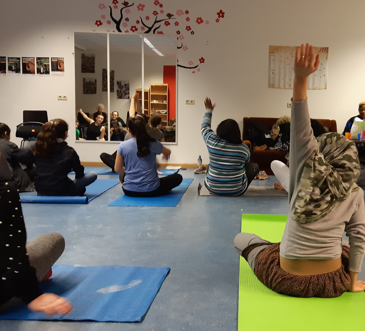 Von hinten sind etwa 5 Frauen zu sehen, die auf Yoga-Matten auf dem Boden sitzen. Zwei strecken gerade weit ihre Arme nach oben. Vor ihnen an der Wand ist ein großer Spiegel, in dem sich noch weitere Frauen im Hintergrund spiegeln.