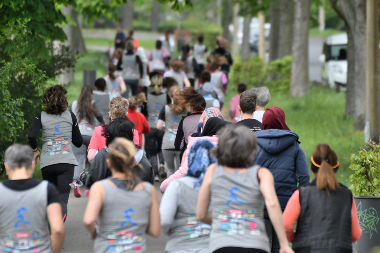 Etwa 40 Frauen rennen in einem begrünten Umfeld alle in eine Richtung. Es sind nur ihre Rücken und Hinterköpfe zu erkennen. Viele von den Läuferinnen tragen ein graues Tanktop mit einer großen blauen Läuferin abgebildet, so wie vielen kleinen Logos.