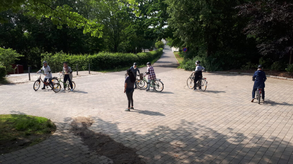 Aus der Ferne sind 6 Frauen mit Fahrrädern und Fahrradhelm fotografiert, die alle jeweils über ihrem Fahrrad stehen, während ihre Füße noch den Boden berühren. In der Mitte des gepflasterten Platzes im Grünen steht eine schwarz gekleidete Frau.
