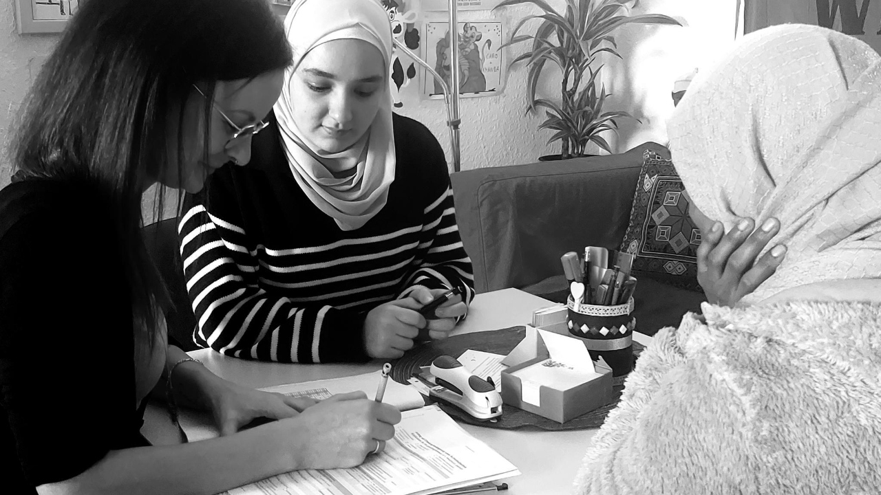 Die schwarz-weiße Aufnahme zeigt drei Frauen an einem quadratischem Tisch. Die linkssitzende Frau mit langen dunklen Haaren füllt gerade ein Formular aus, unter dem eine dicke Akte liegt. Die Frau links von ihr, die ein weißes Kopftuch trägt, schaut auch auf das Antragsformular. Die dritte Frau, die ebenfalls ein Kopftuch trägt, ist nur leicht von hinten zu sehen.
