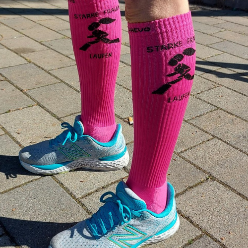 Zwei Unterschenkel in hellblauen Sportschuhen präsentieren die knall pinken Laufsocken des Frauenlaufs. Darauf ist eine gezeichnete Frau im Rennen abgebildet sowie der Spruch "Starke Frauen Laufen".