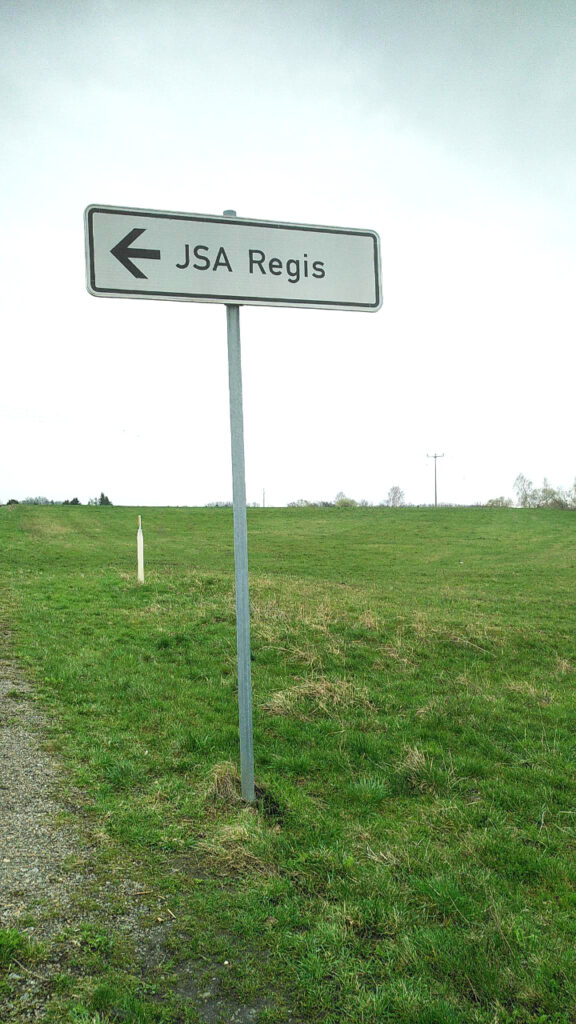 Auf einer grünen Wiese, vermutlich an einem Straßenrand, steht ein graues Schild auf dem mit schwarz "JSA Regis" geschrieben sowie ein dicker Pfeil nach links gezeichnet ist. Der Hintergrund ist sehr hell. Einige Baumspitzen zeichnen sich im Hintergrund ab.