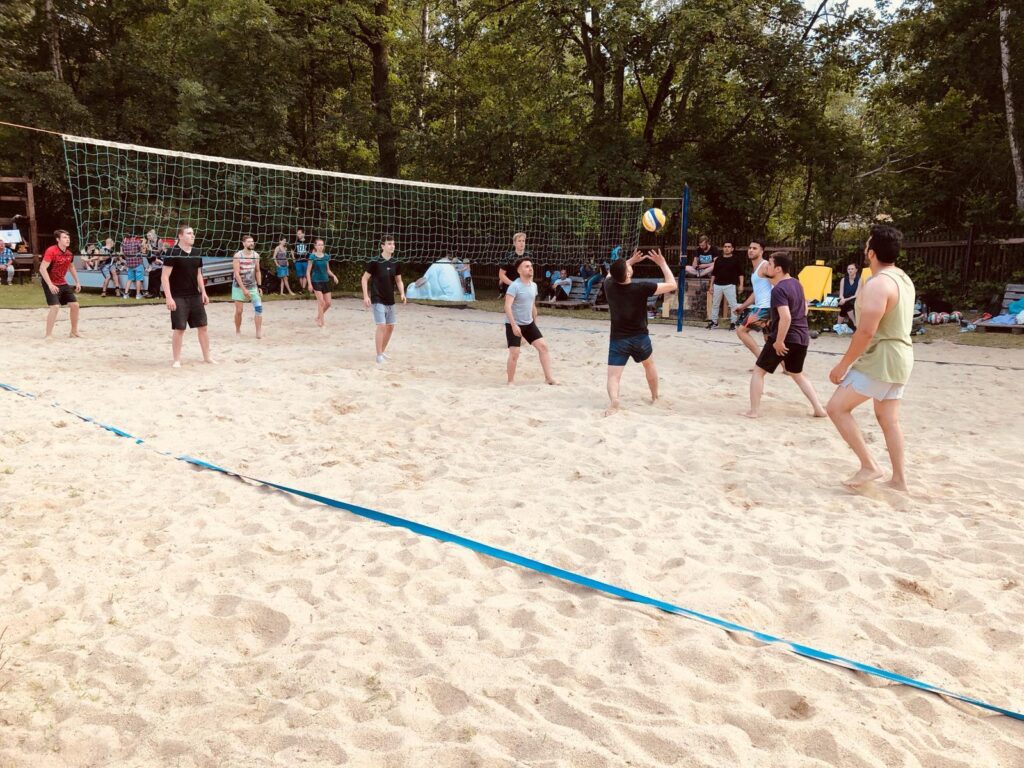 Auf einem Beachvolleyballfeld spielen ungefähr zwölf Personen zusammen Volleyball. Die Personen tragen kurze Hosen und keine Schuhe und mehrere Zuschauer und Zuschauerinnen stehen um das Spielfeld, um den Spiel zuzugucken. Eine Person ist gerade dabei, den Volleyball anzunehmen und einer anderen Person zuzuspielen.