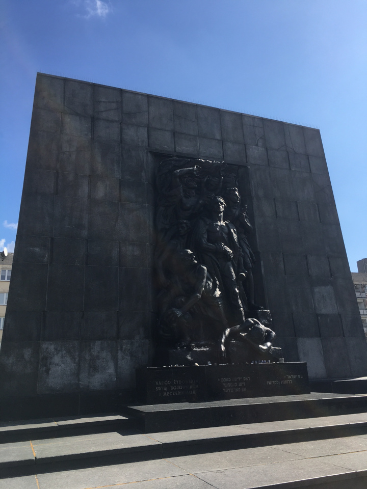 Das Bild zeigt das Warschauer Ghetto-Ehrenmal in Gedenken an den Aufstand im Warschauer Ghetto 1943. Das Denkmal besteht aus großen grauen quadratischen Steinblöcken, die eine Skulpturengruppe umrahmen. Die Skulpturengruppe zeigt exemplarisch Männer unterschiedlichen Alters, die am Warschauer Ghettoaufstand teilgenommen haben.