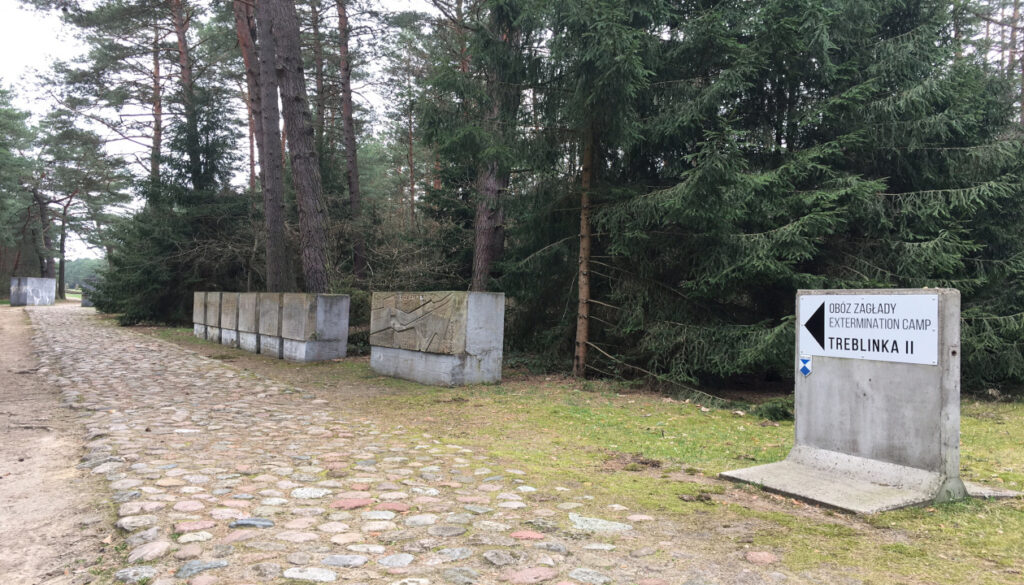Das Bild zeigt den Eingangsbereich der Gedenkstätte Treblinka. Dieser führt über einen Kopfsteinpflasterweg zwischen Bäumen entlang durch zwei große Betonquader. Diese Quader sollen das ursprüngliche Eingangstor des Lagers verdeutlichen.