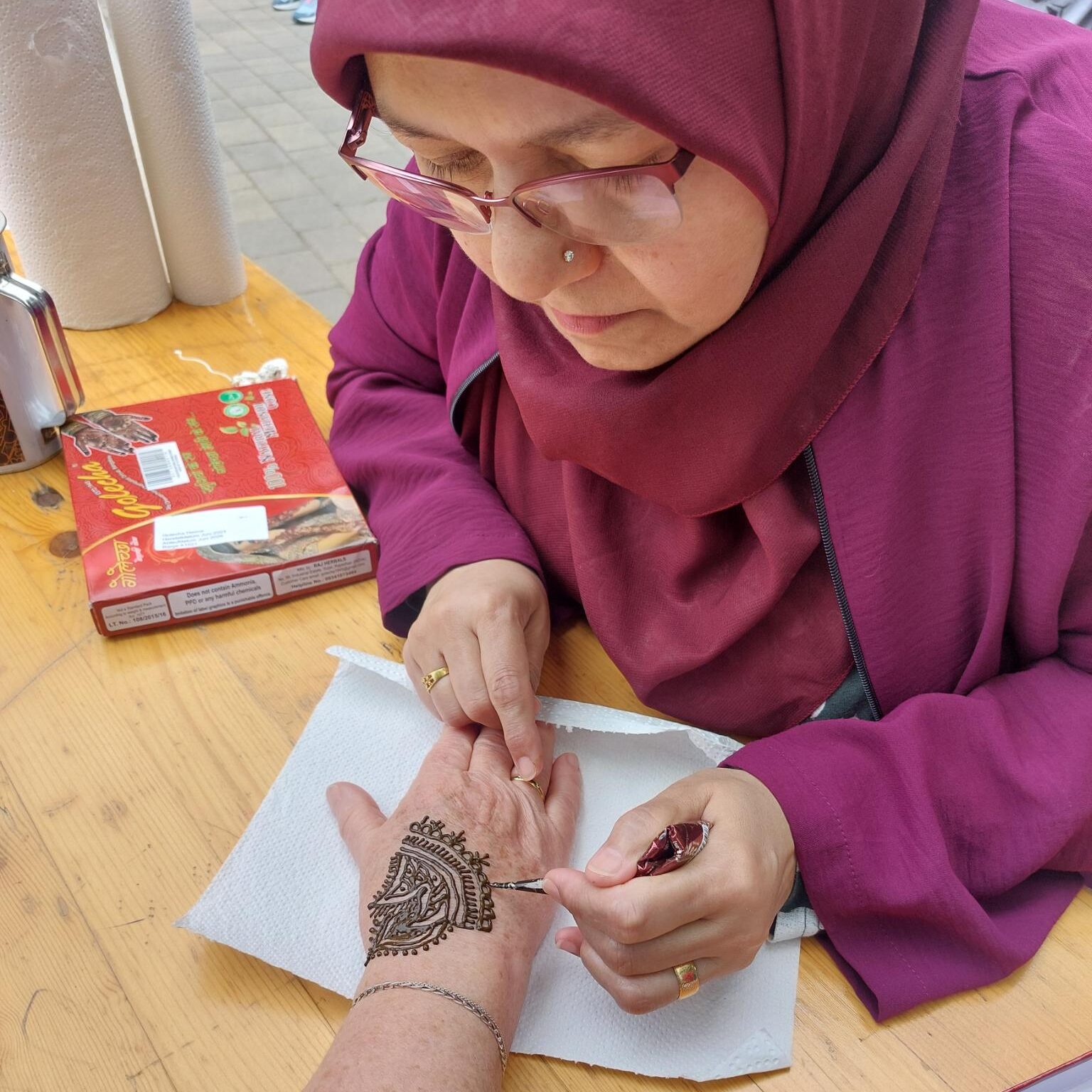 Konzentriert arbeitet eine Frau an einem Henna, dass sie gerade einer anderen Person auf die linke Hand aufträgt. Das Henna-Muster ist sehr detaillreich.