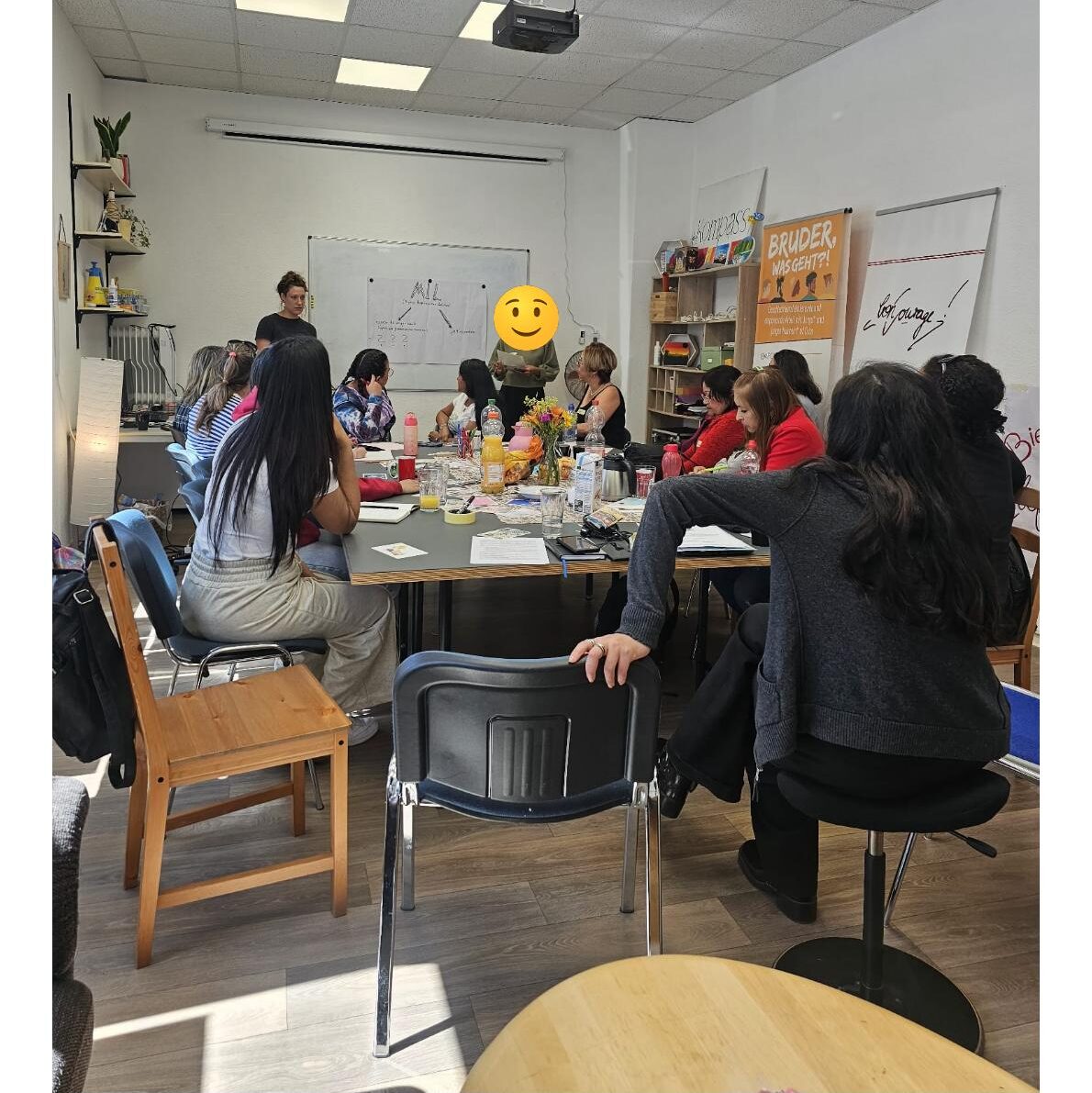 In einem großen Raum sitzen um einen großen Tisch verteilt verschiedene weibliche Personen, deren Blicke bzw. Köpfe den zwei Personen zugewandt sind, die weiter vorn an einem Whiteboard stehen. Auf dem Tisch stehen Getränke und viele haben Notizblöcke vor sich liegen.