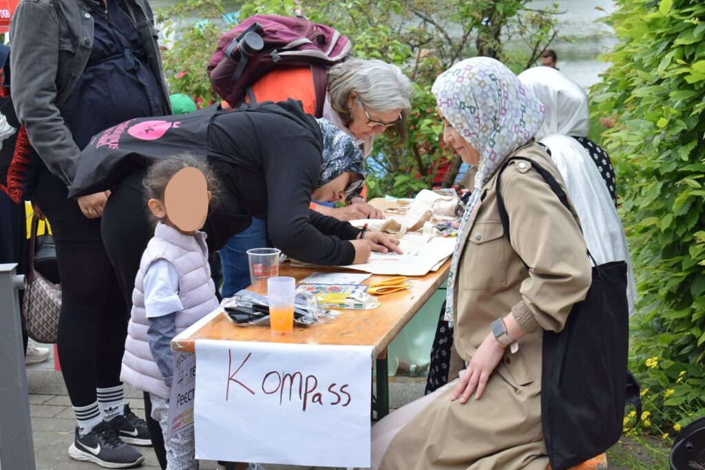 An einer Bierbank, an dessen einer Stirnseite ein handgeschriebener Zettel mit der Aufschrift "Kompass" hängt, sitzen auf der einen Seite zwei Frauen, die beide ein Koopftuch tragen. Auf der anderen Seite sind zum Tisch heruntergebeugt zwei Frauen, die sehr vertieft etwas mit Stiften in der Hand machen.