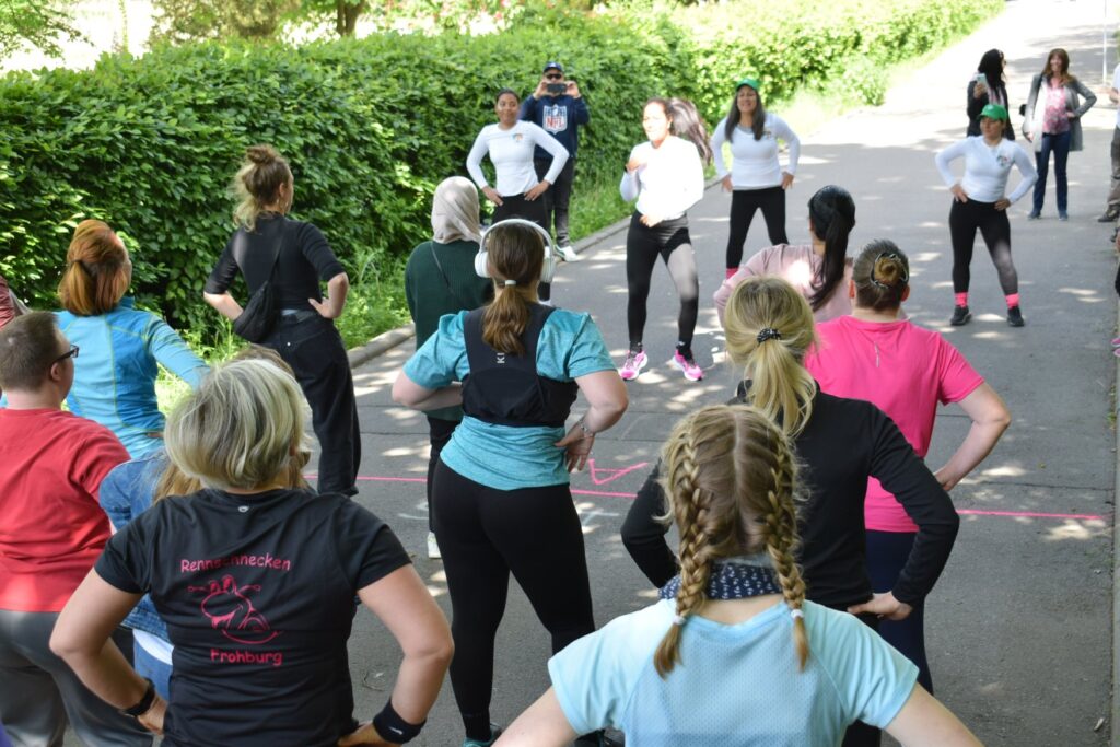 Verschiedene weiblich gelesene Personen stehen auf einer klenen Straße mit in der Hüfte verschränkten Armen und blicken auf vier weitere Frauen in weißen Shirts die vor ihnen sportliche Bewegungen zum Nachmachen ausführen.