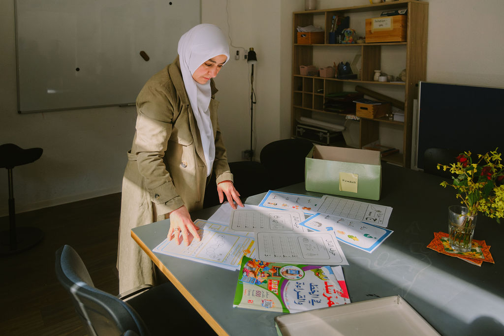 Vor einer Frau mit weißem Kopftuch und beigen Mantel liegen ausgebreitet auf einem Tisch verschiedene Blätter und Hefter, auf denen zum Teil arabische Schriftzeichen erkennbar sind. Im Hintergrund ist ein Whiteboard und ein Schrank mit vielen Materialien.