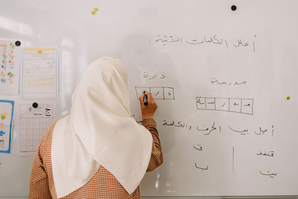 Vor einem Whiteboard steht eine weiblich gelesene Person mit dem Rücken zur Kamera. Mit dem schwarzen Stift in der rechten Hand schreibt sie in Vierecke arabsiche Schriftzeichen. Sie trägt ein weißes Kopftuch und darunter eine karierte Bluse. An dem Whiteboard sind weitere arabische Buchstaben und Wörter geschrieben.