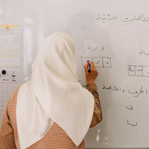 Vor einem Whiteboard steht eine weiblich gelesene Person mit dem Rücken zur Kamera. Mit dem schwarzen Stift in der rechten Hand schreibt sie in Vierecke arabsiche Schriftzeichen. Sie trägt ein weißes Kopftuch und darunter eine karierte Bluse. An dem Whiteboard sind weitere arabische Buchstaben und Wörter geschrieben.