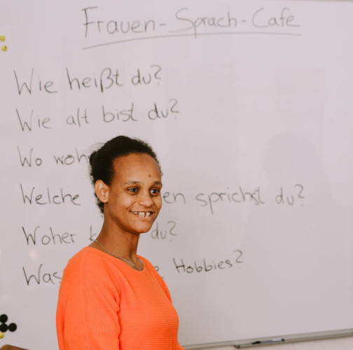 Eine Frau in einem knallig orangenen Pulli steht lächelnd vor einem Whiteboard, mit den Blick in den Raum gewandt. An dem Whiteboard stehen unter der Überschrift Frauen-Sprach-Café verschiedene Kennenlernfragen, zum Beispiel: Wie heißt du? Wie alt bist du? Die anderen lassen sich nur erahnen, da die Frau davorsteht.