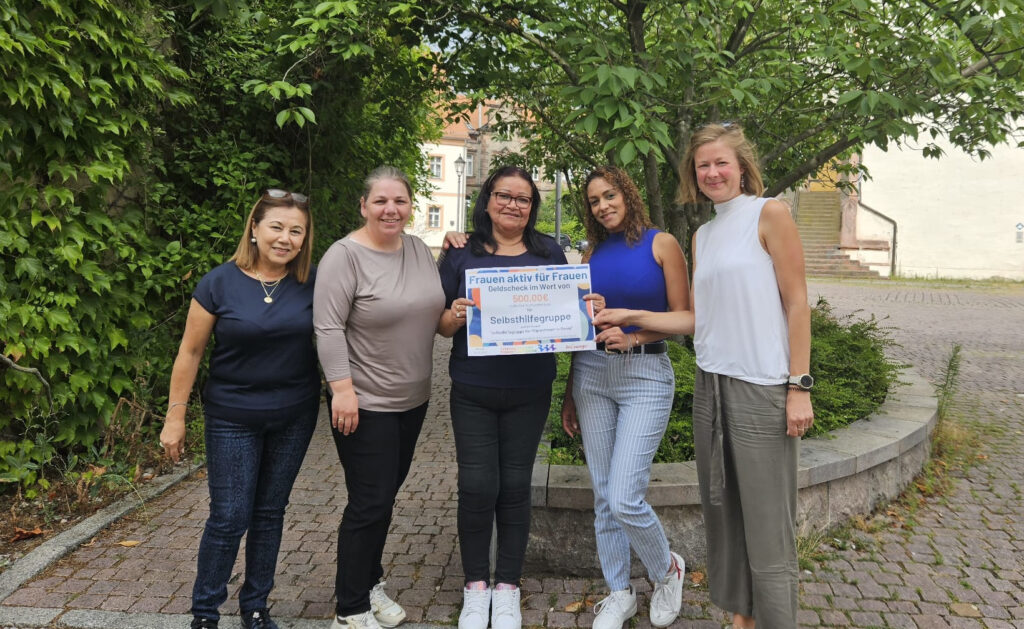 Fünf weiblich gelesene Personen stehen aufgereiht und blicken direkt in die Kamera. Die Stimmung wirkt sommerlich und freudig. Die mittleren Personen halten ein großes Papier in die Kamera, auf dem mitunter "Frauen aktiv für Frauen" und "500€" zu lesen ist.