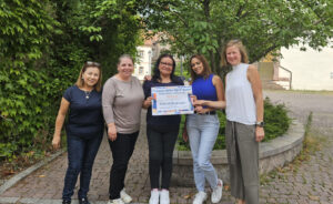 Fünf weiblich gelesene Personen stehen aufgereiht und blicken direkt in die Kamera. Die Stimmung wirkt sommerlich und freudig. Die mittleren Personen halten ein großes Papier in die Kamera, auf dem mitunter "Frauen aktiv für Frauen" und "500€" zu lesen ist.