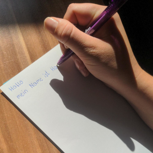 Deutschkurs In einem starken Sonnen-Schattenkontrast zeigt das Bild eine Hand, die einen Kuli-Stift hält und auf einem weißen Blatt Papier die Worte "Hallo, mein Name ist Ha..." verfasst hat.