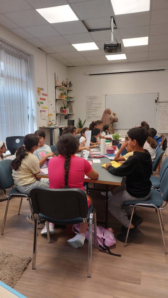 Ungefähr 15 Kinder sitzen zusammen um einen Tisch. Im HIntergrund ist eine Whiteboardtafel abgebildet, an der eine Lehrerin gerade schreibt.