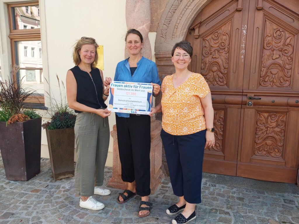 Drei weiblich gelesene Personen stehen vor einem Gebäude mit einer großen hölzernen Eingangstür und halten lächeln einen Papier zur Kamara, bei dem es sich gemäß der Aufschrift um einen Geldcheck in Wert von 121,38€ im Rahmen des Projektes Frauen aktiv für Frauen handelt.