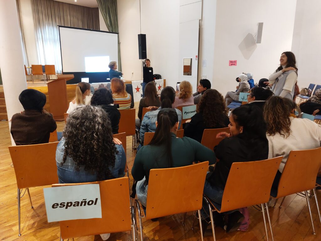 In einen Saal hinein sind von hinten weiblich gelesene Personen zu sehen, die in mehreren Reihen auf Stühlen sitzen, während vor ihnen zwei Frauen gerade etwas aufführen.