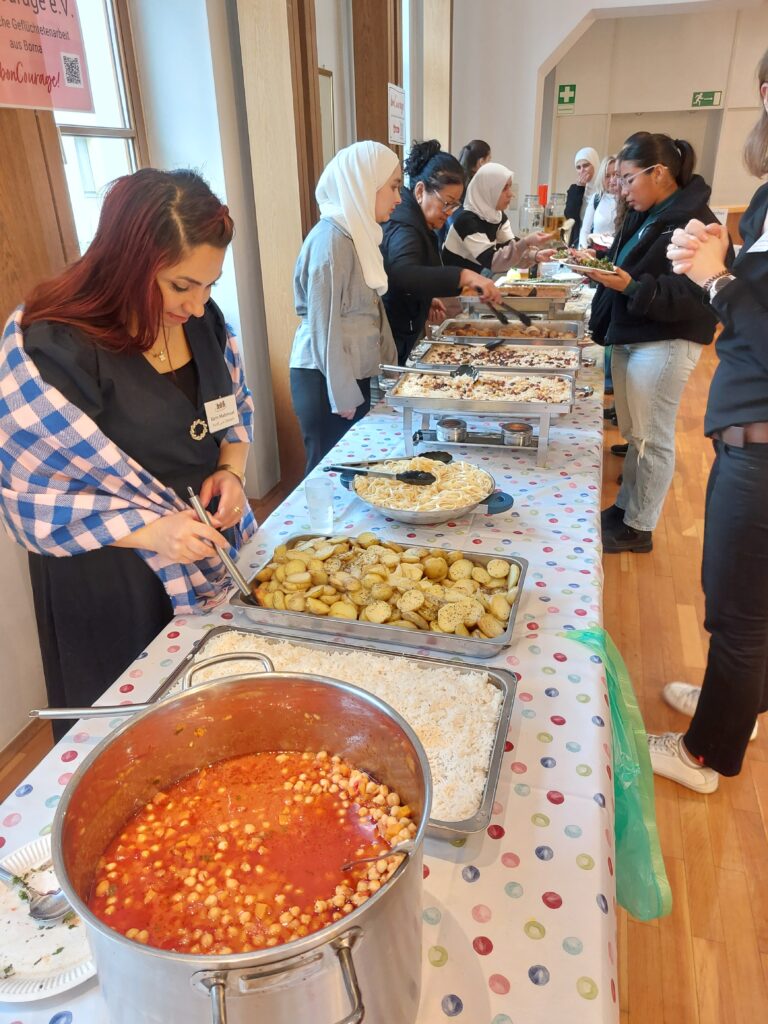 Eine lange Tischtafel ist gedeckt mit reichhaltigem Essen. Auf beiden Seiten stehen verteilt weiblich gelesene Personen, die tels mit dem Essen beschäftigt sind, teils miteinander agieren.