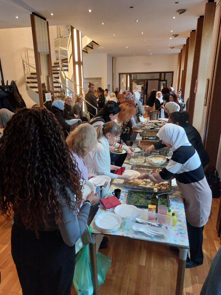 In einem großen Raum sind zahlreich weiblich gelesene Person verteilt. Im Zentrum des Raumes ist eine lange Tischtafel, die gedeckt ist mit verschiedenen Essensplatten. Links davon verteilen Frauen essen an die rechts davon stehenden Frauen. Es herrscht eine trubelige Stimmung.