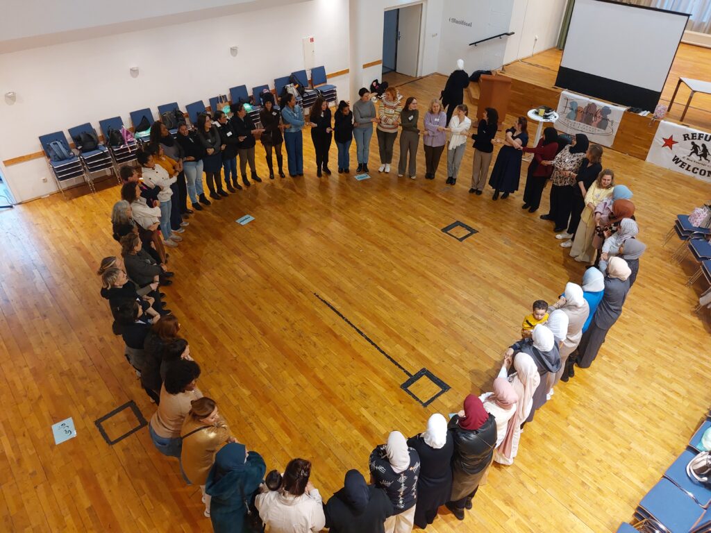 Von oben ist ein Kreis mit rund 60 rein weiblich gelesenen Personen fotografiert, die sich alle über kreuz an den Händen halten und sehr miteinander verbunden wirken.