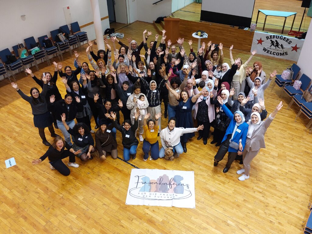 Zahlreiche weiblich gelesene Personen strecken ihre Arme in die Höhe und blicken in Richtung der Kamera. Die Stimmung wirkt ausgelassen und freudig. Vor den Frauen liegt ein Banner mit der Aufschrift: "Frauenkonferenz. Für die Frauen der Zukunft."