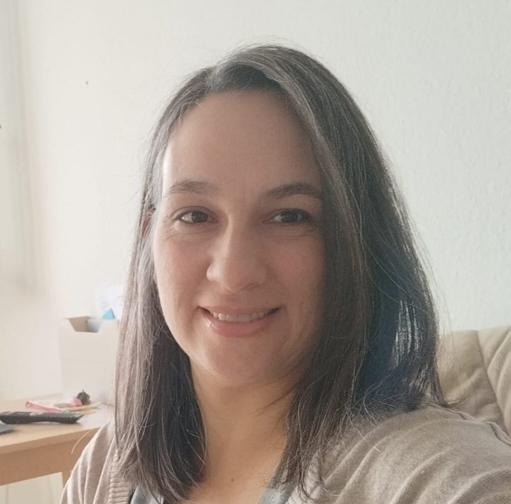 Das Porträt zeigt eine weibliche gelesene Person, die lächelnd in die Kamera blickt. Ihr schulterlanges Haar trägt sie offen und mit einem beigen Pullover ist sie schlicht gekleidet.