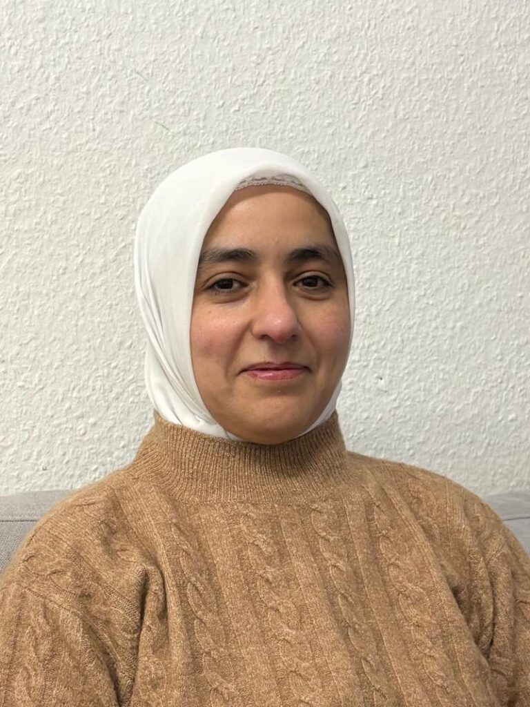 Das Porträt zeigt eine weibliche gelesene Person vor einer weißen Wand, die leicht in die Kamera lächelt und mit einem beigen Rollkragenpullover und einem weißen Kopftuch schlicht gekleidet ist.