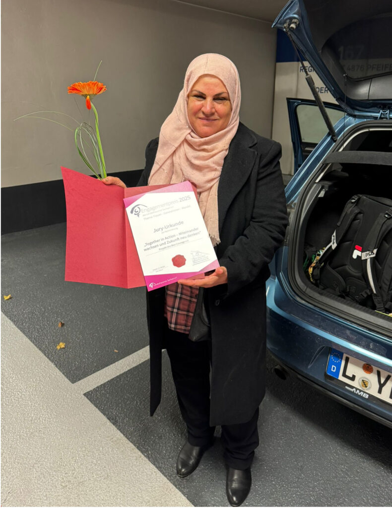 Eine weiblich gelesene Person, elegant mit einem schwarzen Mantel und einem rosa Kopftuch begleitet, hält in die Kamera eine Urkunde. Außerdem hält sie noch ein hübsches Blümchen in der Hand.