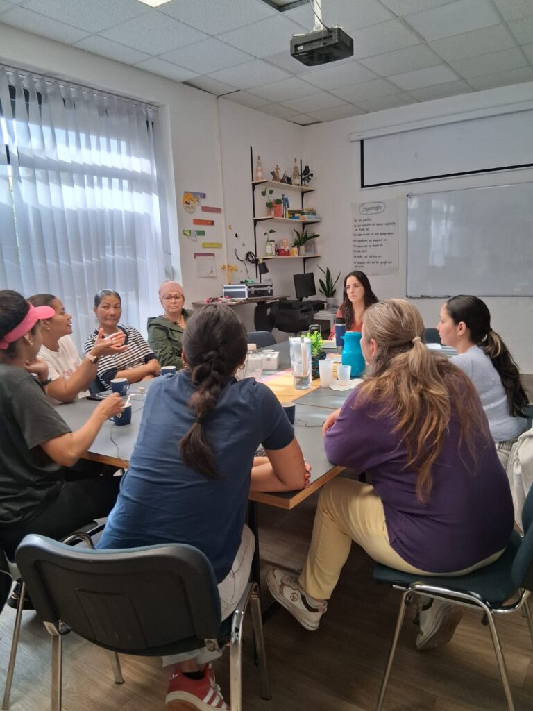 An einem Tisch in einem Raum verteilt sitzen 8 weiblich gelesene Personen. Die aufmerksamen Blicke der meisten sind auf eine von ihnen gerichtet, die in dem Moment zu reden scheint.