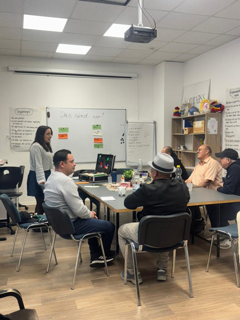 An einem Tisch verteilt sitzen 5 männlich gelesene Personen und eine weiblich gelesene Person steht. Auf dem Tisch stehen verschiedene Dinge, unter anderen ein Moderationskoffer und Getränke. An dem Whiteboard im HIntergrund steht geschrieben "Das sind wir!".