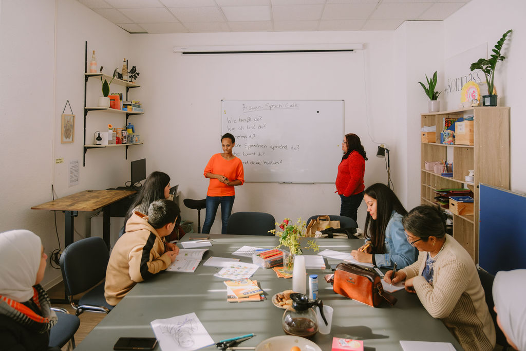 Das Bild wurde während des Frauensprachcafes fotografiert. Man sieht einen großen Tisch an dem konzentrierte Menschen sitzen, die schreiben oder an die Tafel blicken. Man sieht außerdem die Lehrerinnen vor einer Tafel stehen. Auf der Tafel stehen Deutsche Sätze wie z.B. „Wie heißt du?“ „Wie alt bist du?“ etc