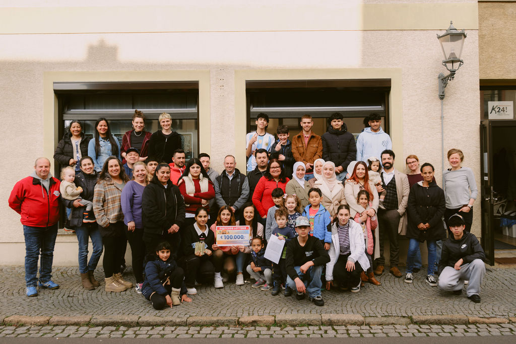 Ein Gruppenbild vor dem K24! Man sieht viele verschiedene Menschen die sich im K24! organisieren und engagieren. Die Menschen hinten stehen und vorne knien und halten ein Spendencheck von der Postcode Lotterie in der Hand.