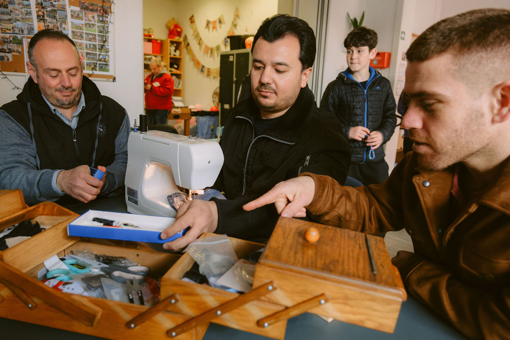 Das Bild wurde während des Nähcafes im K24! fotografiert. Man sieht drei Männer und einen Jungen im Hintergrund. Die Männer sind im Vordergrund des Bildes, unterhalten sich und sind an einer Nähmaschine.