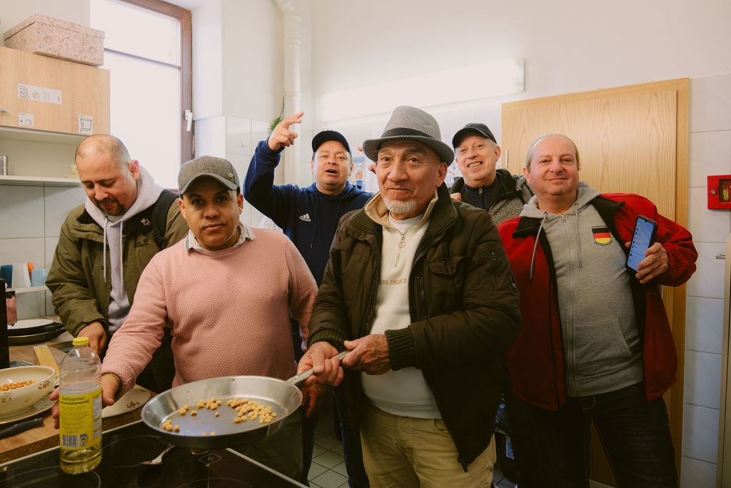 Das Bild zeigt einige Personen der Gruppe „ExodVen“ in der Küche des K24! Beim gemeinsamen Kochen. Die Männer lachen und rufen in Richtung Kamera und die Person vorne im Bild, hält eine große Pfanne in der Hand in der Kichererbsen gebraten werden.