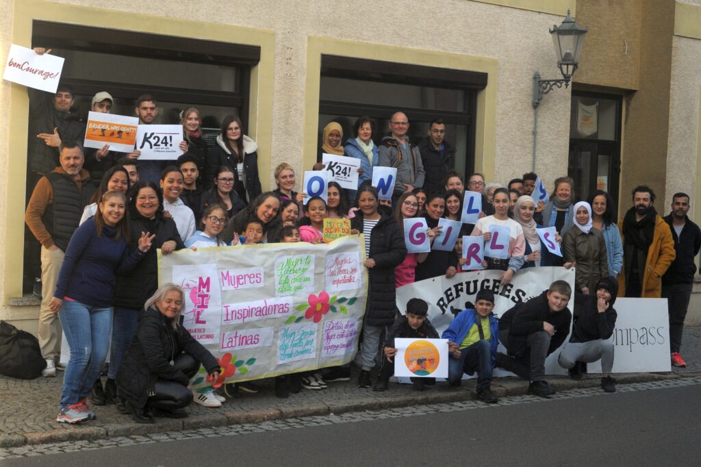 Das Bild zeigt eine Gruppe von Menschen die im K24 engagiert sind. Sie stehen vor den Räumlichkeiten des K24! und halten Schilder in den Händen mit der Aufschrift: „K24!“ – „Kompass“ „Girls“ „Mujeres Inspiradoras Latinas“.