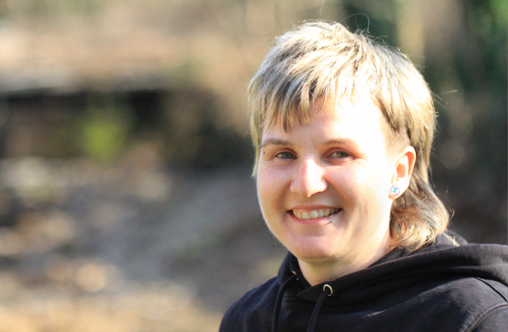 Das Porträt zeigt eine junge, weiblich gelesene Person mit kurz geschnittenen blonden Haaren, die hinten etwas länger sind. Sie lächelt direkt in die Kamera und wird von der Sonne angeleuchtet. In der Lippe und Augenbraue hat sie ein Piercing.