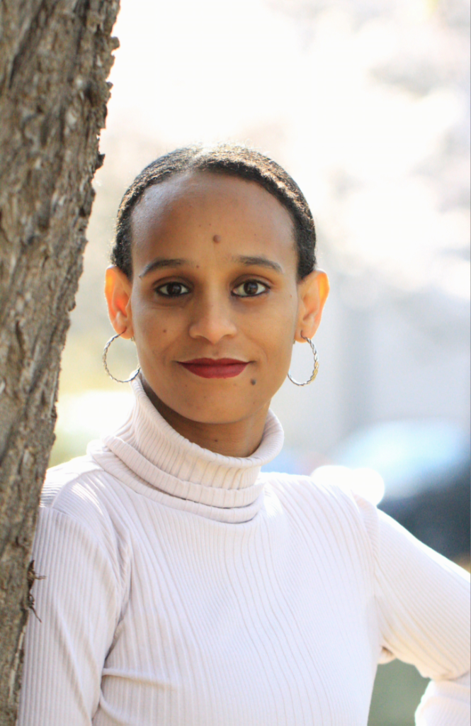 Das Porträt zeigt eine junge Frau mit offenen Blick in die Kamera und einem leichten Lächeln auf den Lippen. Sie lehnt an einem Baumstamm und wirkt sehr selbstbewusst. Sie trägt einen weißen Rollkragenpullover sowie große Griolen.