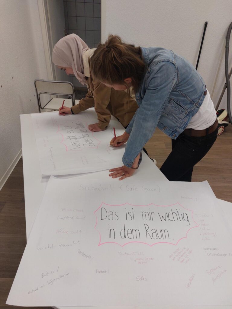 Man sieht 2 Personen die auf Plakate malen. Im Vordergrund ist ein Plakat zu sehen auf dem steht: Das ist mir wichtig in dem Raum.