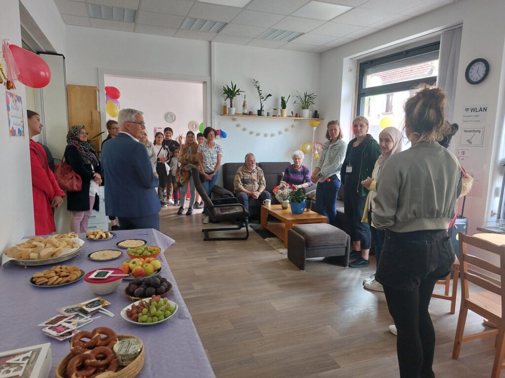 Tag der offenen Tür im K24!. Auf dem Bild sieht man das neu eingerichtete K24! Im Vordergrund ist ein Buffet mit Essen zu sehen, Im Hintergrund stehen und sitzen Menschen im Raum verteilt und lauschen einer Rede.