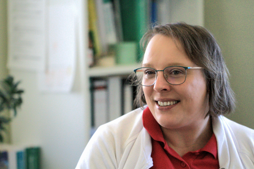 Das Porträt einer weiblich gelesenen Person mittleren Alters, kürzeren Haaren und einer Brille, lächelt leicht an der Kamera vorbei. Sie trägt ein rotes Polo-Short und eine weiße Jacke drüber. Sehr verschwommen sind im Hintergrund Bücher, Aktenordner und Zettel zu erkennen.