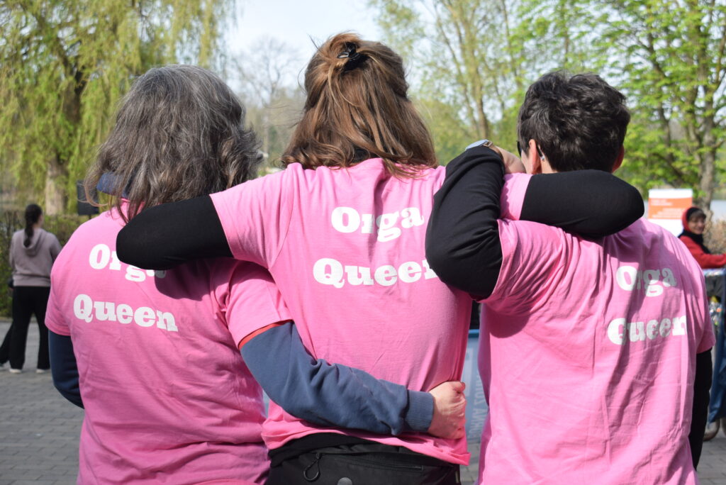 Drei von hinten fotografierte weiblich gelesene Personen mit gleichen pinken Shirts mit der Aufschrift "orga Queen" haben sich in einander verhakt. Das Bild strahlt Kraft und Zusammenhalt aus.